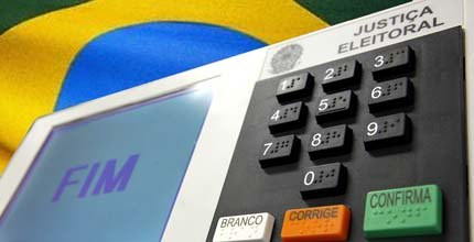 Imagem da urna eletrônica com a bandeira do Brasil atrás.